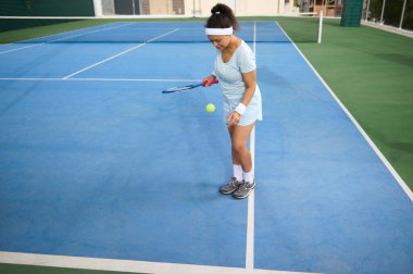 Tenis kıyafeti giymiş bir kadın mavi bir spor kortunda duruyor ve hareketlerine odaklanıyor. Güneşli açık hava ortamı aktif ve odaklanmış bir fitness ortamı sunar.
