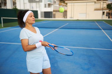 Atletik giyinmiş bir kadın açık hava mavi kortunda güneşli hava koşullarında tenis oynuyor. Odaklanmış ve hazır görünüyor..