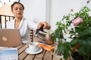 Odaklanmış bir kadın, çiçek açan bir bitkinin yanına oturmuş kahve koyarken dizüstü bilgisayarıyla çalışıyor. Sahne, verimlilik ve sakin boş zamanların bir karışımını vurguluyor..