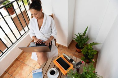 Beyaz cüppeli bir kadın otlarla süslenmiş güneşli bir balkonda otururken uzaktan bilgisayarla çalışıyor. Kahve ve kitap gibi sabah gereksinimleri dengeli bir iş hayatı yaklaşımını yansıtıyor..