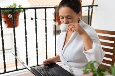 Bir kadın balkonda bornoz giymiş, kahvesini yudumluyor ve laptopunu karıştırıyor. Sahne, açık havada sakin ve verimli bir sabahın özünü yakalıyor..