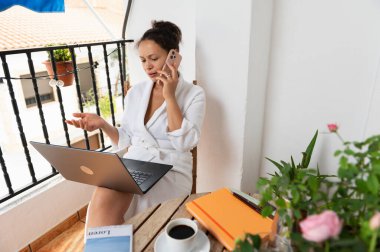 Bir kadın açık hava balkonunda birden fazla iş yapar, rahat bir ortamda dizüstü bilgisayar kullanarak iş görüşmelerini yönetir..