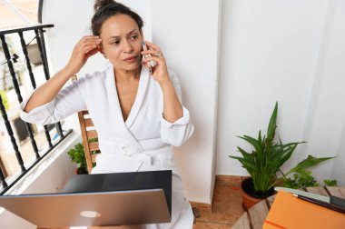 Bir kadın balkonda bornoz giyerek oturuyor, dizüstü bilgisayar kullanırken telefonla konuşuyor. Dekorla çevrili rahat ortam uzaktan çalışma ve iletişim senaryolarını çağrıştırıyor.