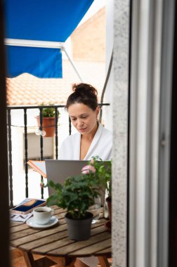 Güzel bitkiler ve çiçeklerle süslenmiş güneşli bir terasta kahvesini yudumlarken dizüstü bilgisayarda çalışan bir kadının huzurlu görüntüsü. Huzurlu ortam rahatlamayı ve odaklanmayı iletir..
