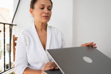 Gündüz giyimli profesyonel bir kadın gündüz bilgisayar kullanırken evde oturur. Atmosfer rahat ve uzak çalışma ya da günlük ev aktivitelerini vurguluyor.