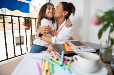Bir anne ve kızı arasındaki neşeli ve tatlı bir bağ anı. Annesi tarafından kucaklanırken kız sıcak bir şekilde gülümsüyor. Çevre, parlak bir balkonda çalışma alanı kurulumunu içeriyor.