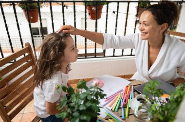 Gülümseyen bir anne ve kızı balkonda bir bağ anının tadını çıkarıyorlar, sanat aktiviteleriyle meşgul oluyorlar. Sahne sıcaklığı, yaratıcılığı ve aile neşesini canlı renklerle yakalıyor, atmosferi güçlendiriyor..