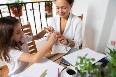 Bir anne ve küçük kızı sıcak bir balkonda otururken yaratıcı bir anı birlikte çizip boyuyorlar. Resim yaratıcılık ve sıcaklıkla hassas bir aile etkileşimi yakalıyor..