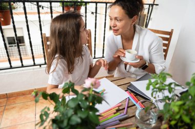 Bir anne ve kızı bir balkonda yaratıcı aktivitelere girişiyor, sanat ve sohbet yoluyla çevresi yeşilliklerle çevrili. Bu ayar yaratıcılığı, rahatlığı ve aile bağlarını iletir.
