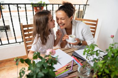 Gülümseyen anne ve kız balkonda yaratıcı aktivitelerin keyfini çıkarıyorlar. Sanat malzemeleri ve saksı bitkileriyle çevrili, aile sevincini ve bağlantısını gösteren samimi bir anı paylaşıyorlar..