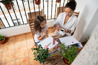 Güneşli bir balkonda çizim yaparken yaratıcı bir bağ kuran bir anne ve kız. Sahne, sakin bir veranda ortamında sanatla uğraşırken sıcaklığı ve şefkati yansıtıyor..