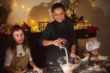 Neşeli bir anne Noel ışıklarının yanında iki çocukla yemek pişirir. Şenlikli bir evde sıcak aile tatili hazırlığı..