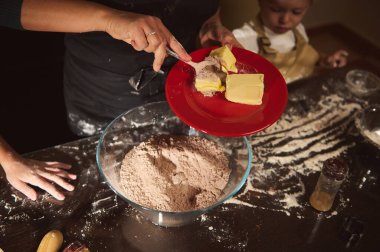 Sıcak bir mutfak sahnesinde bir anne ve çocuk hamur hazırlarken kırmızı tabakta tereyağı tutarken un masanın tozunu alıyor. Sıcak aydınlatma, pişirme etkinliği ve şenlik havası tatil yemekleri ve zencefilli ekmek hazırlama sırasında aile birliğini artırır..