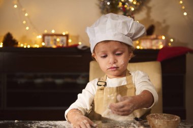 Aşçı şapkası ve önlüğüyle yeni yürümeye başlayan bir çocuk sıcak, şenlikli bir mutfakta zencefilli ekmek evi pişiriyor. Sıcak Noel ışıkları, çocuk tatilde pişirmeye konsantre olurken, neşeli bir aile anını yakalıyor..