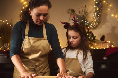 Bir anne ve kızın hamuru yuvarlayıp, dekore edilmiş bir mutfakta zencefilli ekmek evini şekillendirdiği, arka planda yanıp sönen ışıklar ve hediyeler olan sıcak bir tatil anı..
