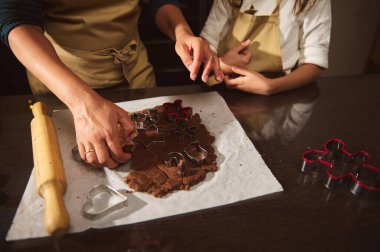 Ailesinin evde zencefilli kurabiye pişirdiği sevgi dolu bir sahne. Eller hamura basar, metal kesiciler hazır ve yakınlarda merdane var. Sıcak, rahat mutfak, çocuklar el sanatlarına katılırken tatil beklentilerini, yaratıcılığı ve paylaşılan mutluluğu yakalıyor. 