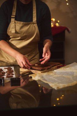 Bej önlüklü bir insan sıcak, şenlikli bir mutfakta tahta bir tahtada kurabiye düzenler. Sıcak ışıklar ve pişirme havası Noel hazırlıklarını ve ev kutlamalarını çağrıştırıyor..