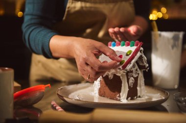 Bir insan sıcak bir mutfakta zencefilli kurabiye evini krema ve renkli şekerlerle süslüyor. Sıcak ışıklar ve şenlik havası tatil yaratıcılığını ve ev kutlamalarını iletir.