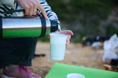Bir el, paslanmaz bir termostan, yeşil bir paspas ve doğal bir zemin ile beyaz bir bardağa, günlük piknik ya da açık hava aktivitesi izlenimi veren sıcak bir sıvı döker..
