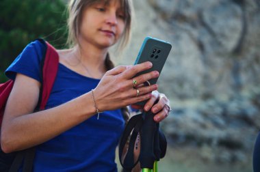 Bir kadın açık havada kayalık bir patikada duruyor, akıllı telefonunu kontrol ederken elinde sırtüstü ve sırt çantasıyla....