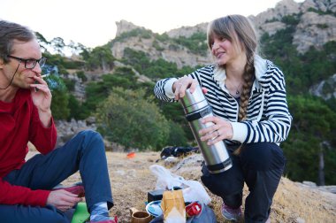 İki yürüyüşçü kayalık bir yamaçta durur, biri termostan su dökerken diğeri arka planda ağaçlar ve engebeli manzarayla sigara içer..