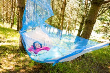 Child girl relaxing on a sunny day in hammock with mosquito net in forest.