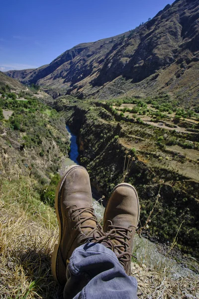 Arid landscape of mountains, mountains, grasslands, and sparse or bushy vegetation with feet and shoes of a tourist in the foreground. South of the Mantaro River Valley.
