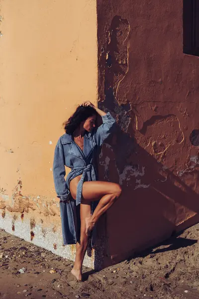 a girl in a denim raincoat poses against the background of a wall. fashion photo shoot on the seashore. stylish shots of a girl model with a beautiful figure.