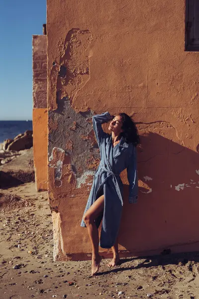 a girl in a denim raincoat poses against the background of a wall. fashion photo shoot on the seashore. stylish shots of a girl model with a beautiful figure.
