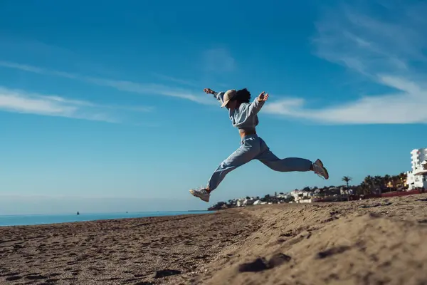 A tanned young woman jumps in a blue suit, a brunette in a tracksuit, an active girl jumps cheerfully on the sea coast. Athletic girl in a tracksuit jumping on the sand