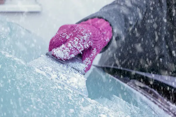 Winter Driving - Close-up of a woman\'s gloved hand using an ice scraper to remove ice from the frozen windscreen of her car before driving.