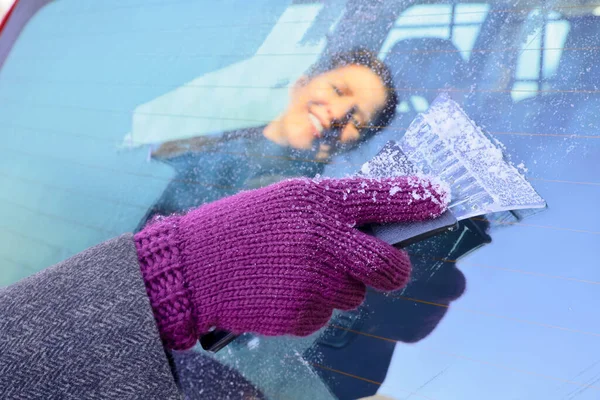 Winter Driving - Close-up of a woman\'s gloved hand using an ice scraper to remove ice from the frozen windscreen of her car before driving.