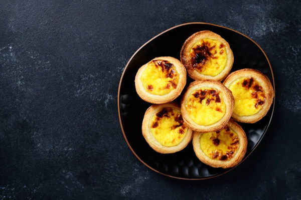 Pastel de nata or Portuguese egg tart. Small tart with a crispy puff pastry crust and a custardy pastry cream filling. Black background, top view