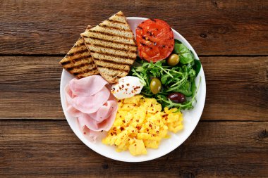 Çırpılmış yumurta, jambon, salata ve kızarmış ekmek ile kahvaltı tabağı. Ahşap arkaplan, üst manzara    