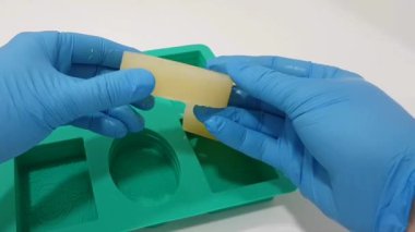 Mavi eldiven giyen ellerin, doğal bir kozmetik laboratuarında silikon kalıp kullanarak yeni yapılmış sabunları öğüttüğü yakın çekim görüntüsü. Bu detaylı görüntüler sanatsal yaratma sürecini vurgular