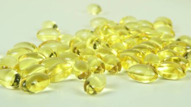 D vitamini içeren şeffaf sarı kapsüllerin yakın görüntüsü, beyaz plastik bir kaptan dökülen, pürüzsüz inci benzeri dokusunu ve makro detay olarak berraklığını gözler önüne seren.