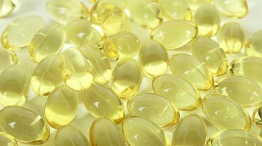 D vitamini içeren şeffaf sarı kapsüllerin yakın görüntüsü, beyaz plastik bir kaptan dökülen, pürüzsüz inci benzeri dokusunu ve makro detay olarak berraklığını gözler önüne seren.