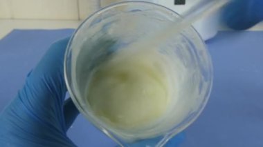 Laboratuvarda hazırlanan kozmetik emülsiyonun makro çekimi. Video, operatörün eldivenli ellerini bir beher kabında karıştırırken yakalıyor. Cilt bakımı, kozmetik ürünleri, laboratuvar süreçleri ve ilaç geliştirme ile ilgili konular için ideal..