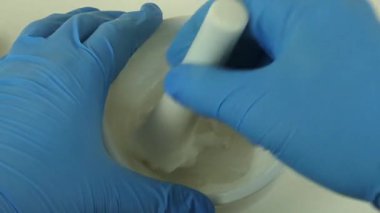 Mavi eldivenli eldivenli ellerin porselen havan ve tokmak kullanarak krem karıştırdığı kozmetik laboratuvar sürecinin makro görüntüsü.. 