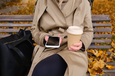 Akıllı telefonu ve bir fincan kahvesi olan bir kız sonbahar parkında bir bankta oturuyor. Bir kadın mesaj atıyor ya da cep telefonunda uygulama kullanıyor. Sıcak bir içecek, kağıt bardakta sıcak bir yürüyüş için.