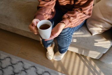 Bir kızın elinde bir fincan çay ya da kahve. Sabahleyin, kız sıcak çay içer. Evin rahatlığının tadını çıkar. Pencereden güneş ışınları alan bir kadının ellerinde beyaz bir fincan.