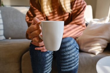 Bir kızın elinde bir fincan çay ya da kahve. Sabahleyin, kız sıcak çay içer. Evin rahatlığının tadını çıkar. Pencereden güneş ışınları alan bir kadının ellerinde beyaz bir fincan.