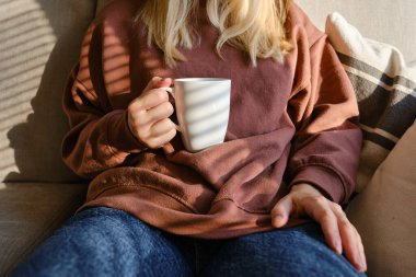 Bir kızın elinde bir fincan çay ya da kahve. Sabahleyin, kız sıcak çay içer. Evin rahatlığının tadını çıkar. Pencereden güneş ışınları alan bir kadının ellerinde beyaz bir fincan.
