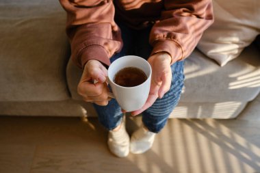 Bir kızın elinde bir fincan çay ya da kahve. Sabahleyin, kız sıcak çay içer. Evin rahatlığının tadını çıkar. Pencereden güneş ışınları alan bir kadının ellerinde beyaz bir fincan.