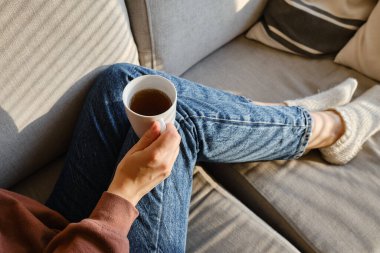 Bir kızın elinde bir fincan çay ya da kahve. Sabahleyin, kız sıcak çay içer. Evin rahatlığının tadını çıkar. Pencereden güneş ışınları alan bir kadının ellerinde beyaz bir fincan.