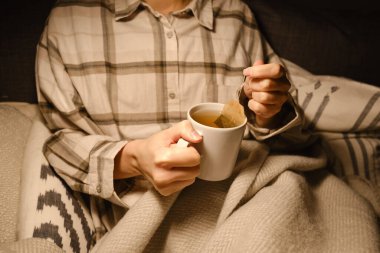 A cup of tea in the hands of a girl. The girl is drinking hot tea. A girl in pajamas is wrapped in a blanket and is drinking delicious tea. Enjoy the comforts of home. White cup close up in woman hand
