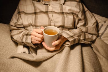 A cup of tea in the hands of a girl. The girl is drinking hot tea. A girl in pajamas is wrapped in a blanket and is drinking delicious tea. Enjoy the comforts of home. White cup close up in woman hand