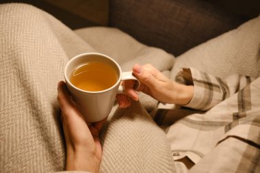 A cup of tea in the hands of a girl. The girl is drinking hot tea. A girl in pajamas is wrapped in a blanket and is drinking delicious tea. Enjoy the comforts of home. White cup close up in woman hand