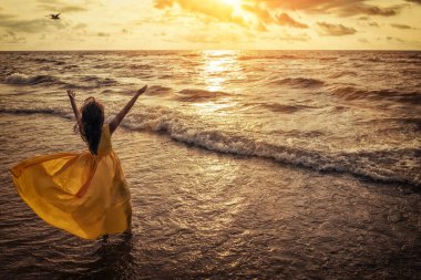 Yazın gün batımında deniz manzarası. Sahilde bir kadın. Elleri havada mutlu genç bir kadın, sarı çırpınan bir elbiseyle deniz kenarında tasasız yürüyor. Yukarıdan görüntüle