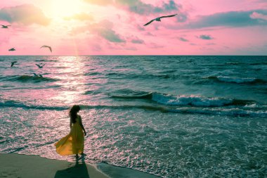 Pembe gündoğumunda deniz manzarası ve güzel bir gökyüzü. Sahildeki kadın. Sarı kanat çırpan elbiseli genç mutlu kadın deniz kıyısında yürüyor. Kız sihirli gün doğumuna bakıyor. Yukarıdan görüntüle.